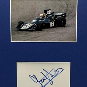 Puede incluir: Fotografía enmarcada de un coche de carreras de Fórmula 1 en movimiento, con el número 3 y logotipos de patrocinadores. Debajo, una firma azul sobre un fondo blanco. El marco es azul oscuro.