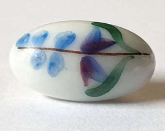 Small oval ceramic button hand painted with blue flowers and green leaves. Porcelain. Circa 1900s. 21/32&quot; by 3/8&quot;. PB143