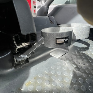 May include: A black metal cup holder with two cup holders and the word "KASPEN" printed on the front. The cup holder is mounted in a car.