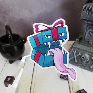May include: A blue and pink gift box sticker with a white bow and a pink tongue sticking out of the box. The box has a menacing expression with white eyes and sharp teeth.