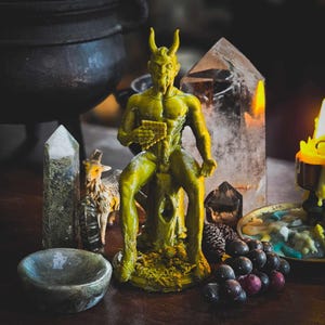 May include: A green figurine of a horned, male figure sitting on a tree stump. The figure is holding a small, rectangular object in its hand. The figurine is surrounded by crystals, a small goat figurine, and grapes.