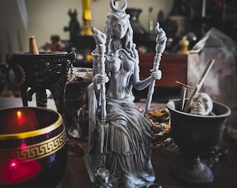 May include: A gray statue of a seated woman with a crescent moon crown, holding two staffs with flames. The statue is surrounded by candles, a skull, and other mystical items, creating a gothic or witchy aesthetic.