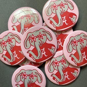 May include: A collection of circular pin-back buttons featuring a light pink background with a gray elephant illustration. The elephant wears red star-shaped glasses and a red shirt with a white letter A printed on the front of the garment.