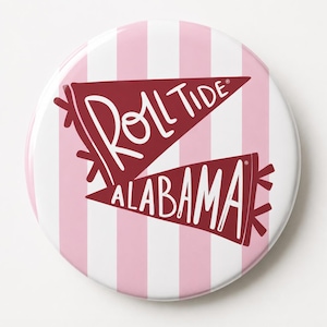 May include: A circular button featuring a background of vertical light pink and white stripes. Two maroon pennants are layered over the stripes, with the white text "ROLL TIDE" on the top pennant and "ALABAMA" on the bottom pennant.