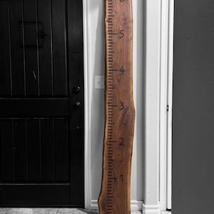 May include: A tall, vertical wooden growth chart made from a single slab of dark brown wood with a natural live edge. The board features black engraved markings and numbers from one to six, indicating height measurements in feet for tracking growth.