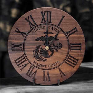 May include: A circular wooden wall clock featuring dark brown wood grain with black engraved Roman numerals. The center displays the United States Marine Corps emblem, including an eagle, globe, and anchor, with black hour and minute hands.