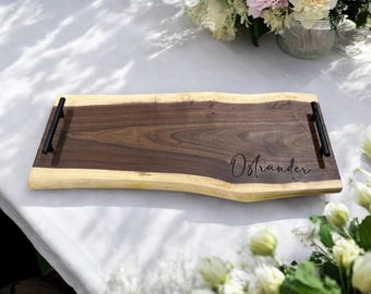 Puede incluir: Una bandeja de madera rectangular con un centro marrón oscuro y un borde de color claro. La bandeja tiene asas negras y el nombre "Ostrander" está grabado. La bandeja está sobre un mantel blanco.