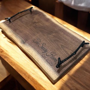 May include: A rectangular wooden serving tray with a natural live edge and two black metal handles shaped like tree branches. The surface is engraved with the text The Sneary Home EST 2025 in a cursive and block font combination.