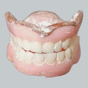 May include: A set of full dentures featuring a pink acrylic base that mimics gum tissue and a full arch of white, realistic-looking prosthetic teeth. The upper and lower plates are joined together, showing the biting surface and structure.