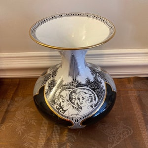 May include: A decorative ceramic vase with a wide, oval opening and a bulbous base. The vase features black and white illustrations of figures and landscapes, with gold trim around the rim and base. The vase is on a brown surface.