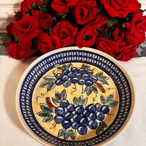 May include: A round ceramic bowl featuring a hand-painted design of two clusters of blue grapes with green leaves on a cream-colored background. The rim is decorated with a dark blue patterned border. The bowl is positioned in front of red roses.