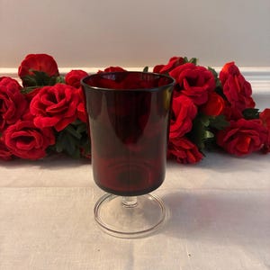May include: A ruby red glass goblet with a clear, circular base. The goblet is cylindrical with a slightly flared rim. Red roses are in the background. The goblet is likely used for serving beverages.