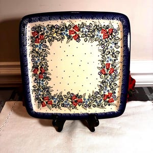 May include: Square ceramic serving dish with a floral design. The dish has a navy blue border with a detailed pattern. The interior features a cream-colored background with a floral arrangement of red roses and blue flowers.