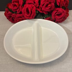 May include: A white oval ceramic serving dish featuring a central divider that splits the interior into two equal sections. The dish has a smooth, glossy finish and a raised rim, providing a clean and functional design for serving various food items.