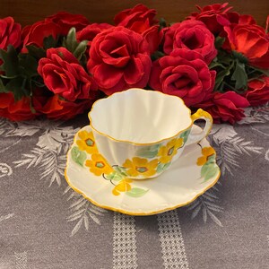 May include: A white teacup and saucer with a yellow and green floral pattern. The cup and saucer have a scalloped edge.