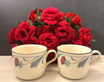 May include: Two cream-colored ceramic mugs featuring a delicate floral pattern with red flowers, blue leaves, and thin blue rim accents. The mugs have a rounded body and a side handle, positioned side by side in front of a cluster of red roses.