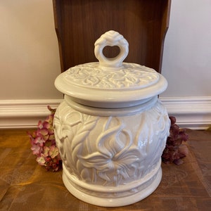 May include: Off-white ceramic jar with lid, featuring embossed floral designs. The jar has a heart-shaped handle on the lid. The jar is round and sits on a table with a brown tablecloth. A wooden shelf is in the background.