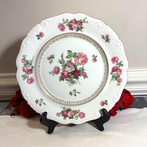 May include: White porcelain plate with a scalloped edge, adorned with pink and red roses and gold trim. The plate is supported by a black stand, showcasing a classic floral design. A decorative piece for serving or display.