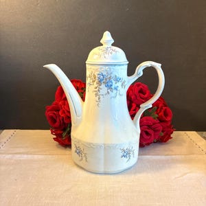 May include: A white porcelain coffee pot with a fluted body and a matching lid. The vessel features delicate blue rose floral patterns with green leaves and brown stems, a curved handle, and a long, slender spout with subtle embossed details.