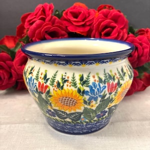 May include: A ceramic planter with a rounded body and a flared rim, featuring a hand-painted floral pattern of yellow sunflowers, blue flowers, and red tulips on a cream background, with a dark blue decorative border along the top and base.