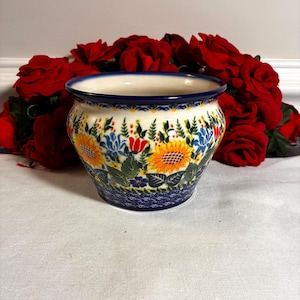 May include: A ceramic flower pot with a blue rim and a floral design featuring sunflowers, blue flowers, and green leaves. The pot has a white background and a blue base. Red roses are in the background.