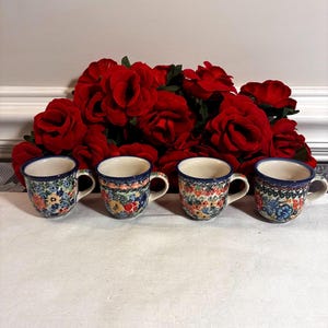 May include: Four ceramic mugs with floral patterns in blue, red, yellow, and green. Each mug has a dark blue rim and handle. The mugs are arranged in front of a backdrop of red roses.