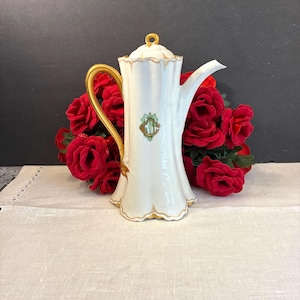 May include: A white porcelain coffee pot with a fluted body, a gold-painted handle, and a gold-trimmed lid. The front features a green and gold decorative crest with the letters 'GC'. The base and rim have scalloped edges with gold accents.