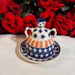 May include: A ceramic butter dish with a lid featuring a doll-shaped handle. The lid is decorated with blue stars on a cream background and orange vertical stripes. The base plate has a matching blue star pattern along the outer rim.