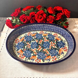 May include: An oval ceramic baking dish featuring a cream-colored interior decorated with blue roses, orange accents, and green leaves. The wide rim displays a dense blue and white geometric pattern. A cluster of red roses sits behind the dish.