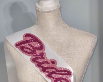 May include: A white satin sash featuring the word Bride in layered, glittery pink and dark magenta script lettering. The sash is displayed on a neutral-toned mannequin torso, showcasing the bold, textured typography against the smooth fabric.