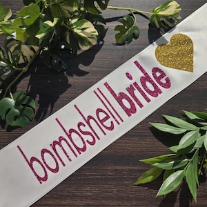 Puede incluir: Una banda de satén blanco con el texto "bombshell bride" en letras de purpurina rosa, acompañada de un icono de corazón de purpurina dorada situado a la derecha del texto. La banda se muestra en diagonal en el encuadre.