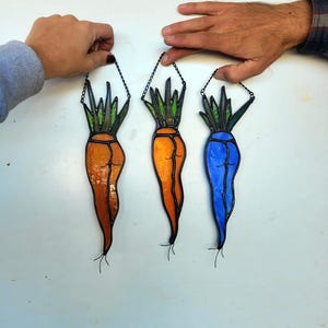 May include: Three stained glass carrot ornaments in orange, orange-yellow, and blue. Each carrot has green and orange-yellow leaves and is suspended by a black chain. The carrots are held up by two hands.