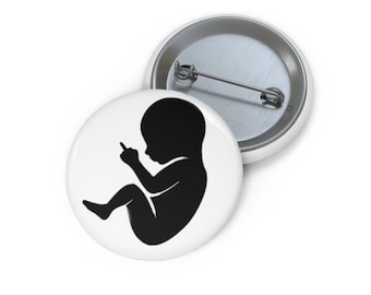 May include: Two white button pins. One pin features a black silhouette of a fetus giving the middle finger. The second pin is partially visible, showing the pin back. The pins are round and have a shiny finish.