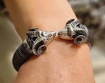 May include: A silver bracelet with two ram heads on the ends. The bracelet is made of a woven metal mesh.
