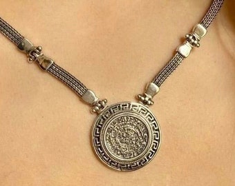 May include: A silver chain necklace with a round pendant featuring an intricate design. The pendant is surrounded by a decorative border.