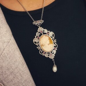 May include: A silver necklace with a cameo pendant featuring a woman's profile. The pendant is surrounded by a delicate filigree design with pink gemstones. A single pearl dangles from the bottom of the pendant.
