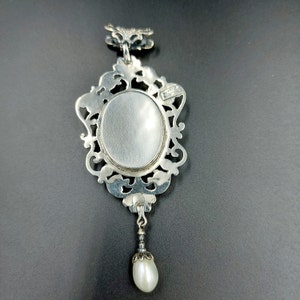 May include: Silver pendant with an ornate frame and a blank oval space for a photo or cameo. A small pearl dangles from the bottom of the pendant.