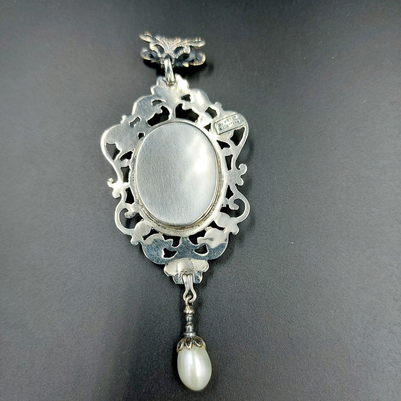 May include: Silver pendant with an ornate frame and a blank oval space for a photo or cameo. A small pearl dangles from the bottom of the pendant.