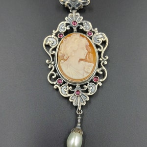 May include: A silver and gold pendant with a cameo of a woman's profile. The pendant is adorned with red gemstones and a pearl drop.