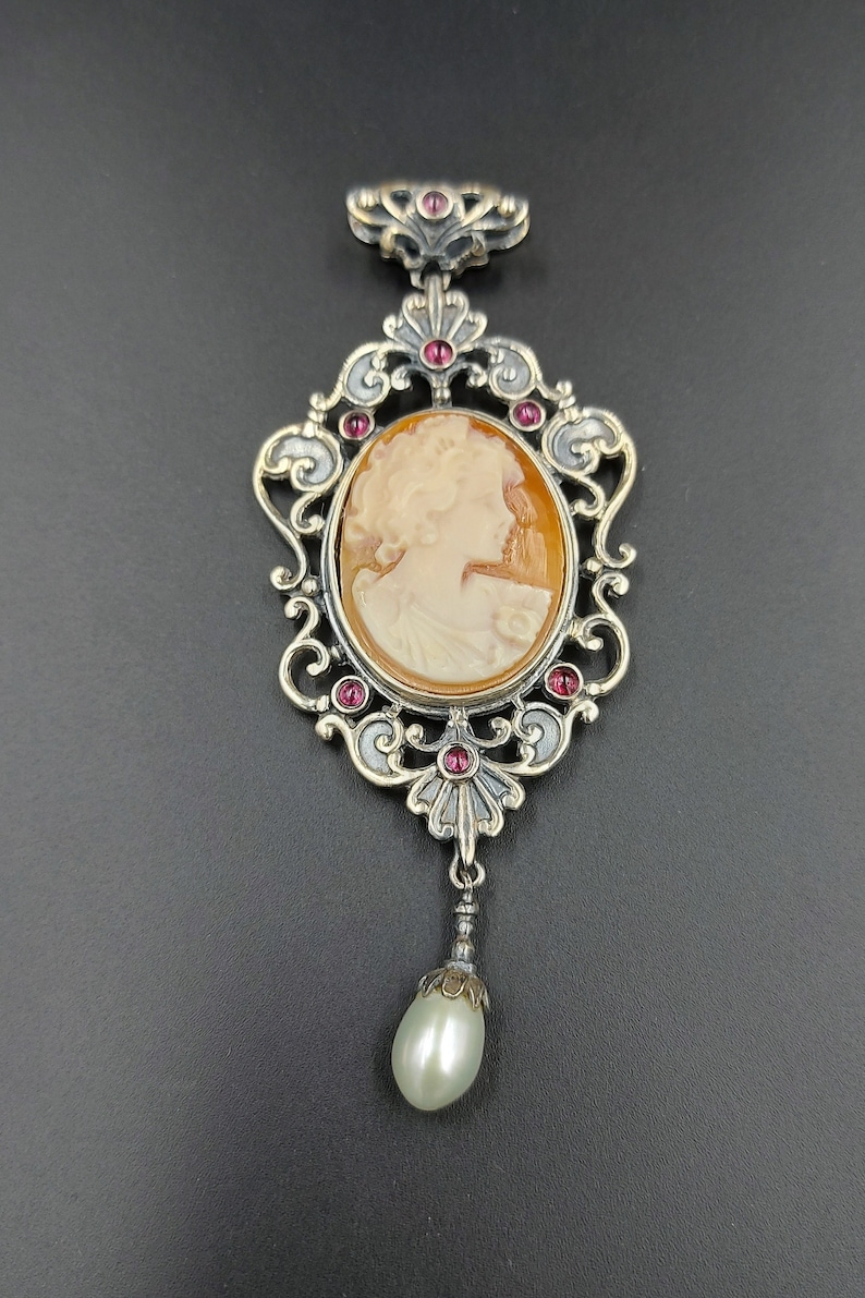 May include: A silver and gold pendant with a cameo of a woman's profile. The pendant is adorned with red gemstones and a pearl drop.