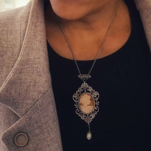 May include: A silver chain necklace with a cameo pendant featuring a woman's profile. The pendant is surrounded by an ornate silver frame with small purple gemstones. A single pearl dangles from the bottom of the pendant.