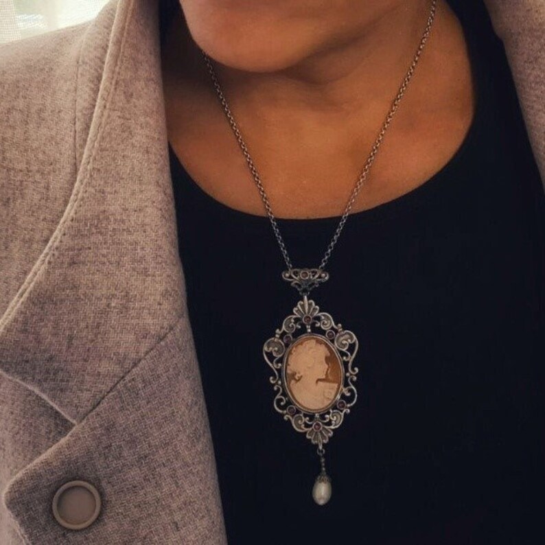 May include: A silver chain necklace with a cameo pendant featuring a woman's profile. The pendant is surrounded by an ornate silver frame with small purple gemstones. A single pearl dangles from the bottom of the pendant.