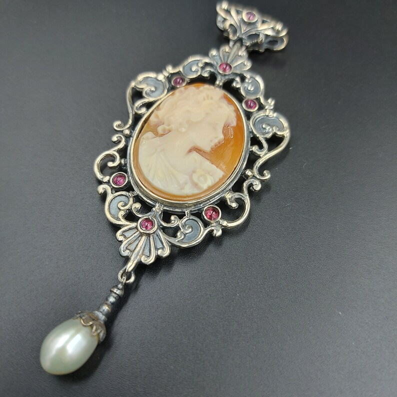 May include: A silver pendant with a cameo of a woman's profile in a delicate, ornate frame. The frame is decorated with small pink gemstones and a pearl drop hangs from the bottom.