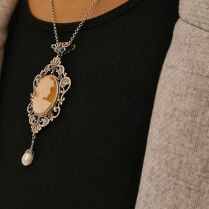 May include: A silver necklace with a cameo pendant featuring a woman's profile. The cameo is surrounded by an ornate silver frame and hangs from a delicate silver chain. A single pearl dangles from the bottom of the pendant.