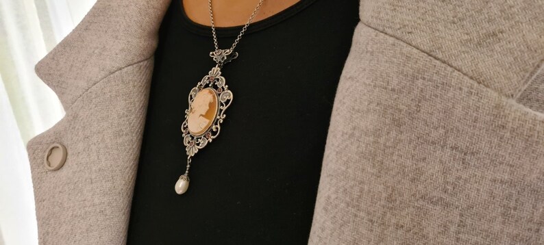 May include: A silver necklace with a cameo pendant featuring a woman's profile. The cameo is surrounded by an ornate silver frame and hangs from a delicate silver chain. A single pearl dangles from the bottom of the pendant.