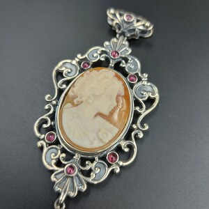 May include: A silver pendant with a cameo of a woman's profile. The cameo is surrounded by a decorative filigree frame with pink gemstones.