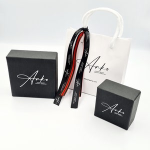May include: Two black gift boxes with the logo "Anko Handmade Greek Jewelry" and the website address "ankojewelry.com" printed on them. A white gift bag with black handles and a red and black ribbon is also shown.