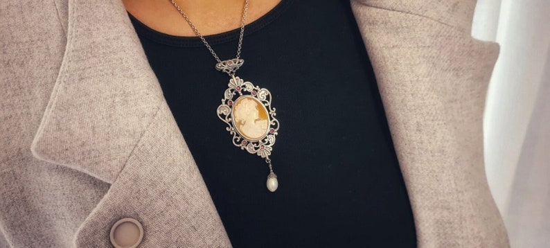May include: A silver necklace with a cameo pendant featuring a woman's profile. The pendant is surrounded by a delicate filigree design with pink gemstones. A single pearl dangles from the bottom of the pendant.