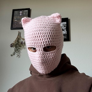 May include: A light pink crochet balaclava with cat ears. The handmade head covering features eye openings and a textured, open weave design. The balaclava is worn over the head and neck, providing warmth and coverage.