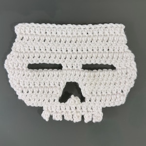 May include: A white, crocheted skull-shaped coaster. The coaster has two horizontal eye slits and a triangular nose opening. The skull design is made with a textured stitch, giving it a handmade appearance. The coaster is likely used for decorative purposes.